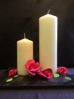 Two large cream candles with red Evolon roses and green Evolon leaves standing on a dark slate tile.