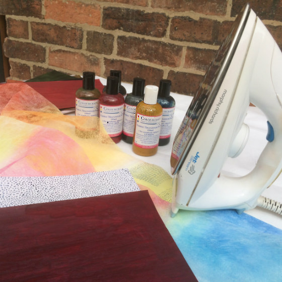 Sheets of A4 paper painted with transfer paints, one solid red and one with a speckled design, bottles of transfer paints, domestic iron and piece of coloured Lutradur.