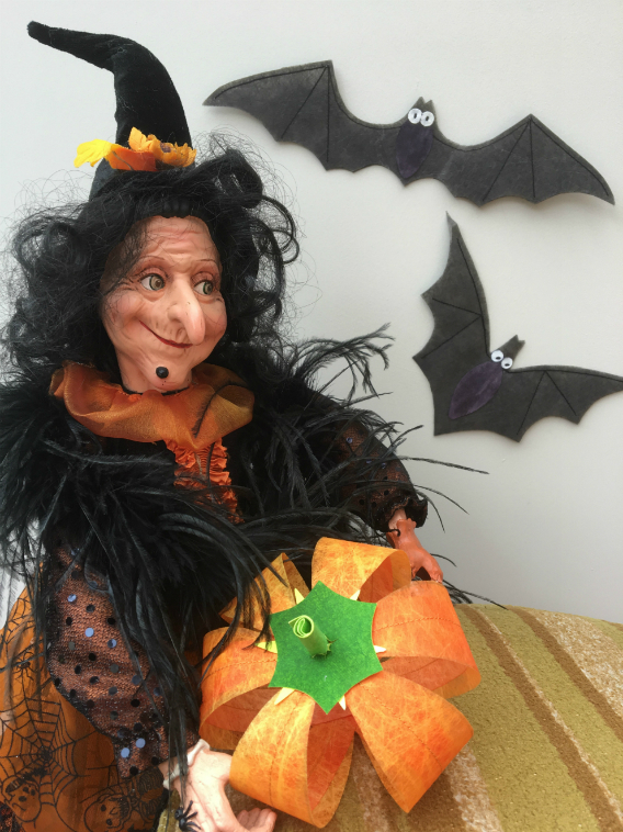 Smiling witch, dressed in orange and black with crooked witch's hat, holding a pumpkin made out of Lutradur. Behind her are two bats made out of Lutradur Black.