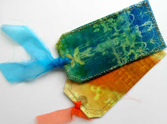 Two rectangular gift tags one yellow and orange, the other blue and green. The blue tag is decorated with a printed green chandelier and has machine stitching around the outer edge. The other tag has been printed with large text, but the writing is decorative and not legible. 