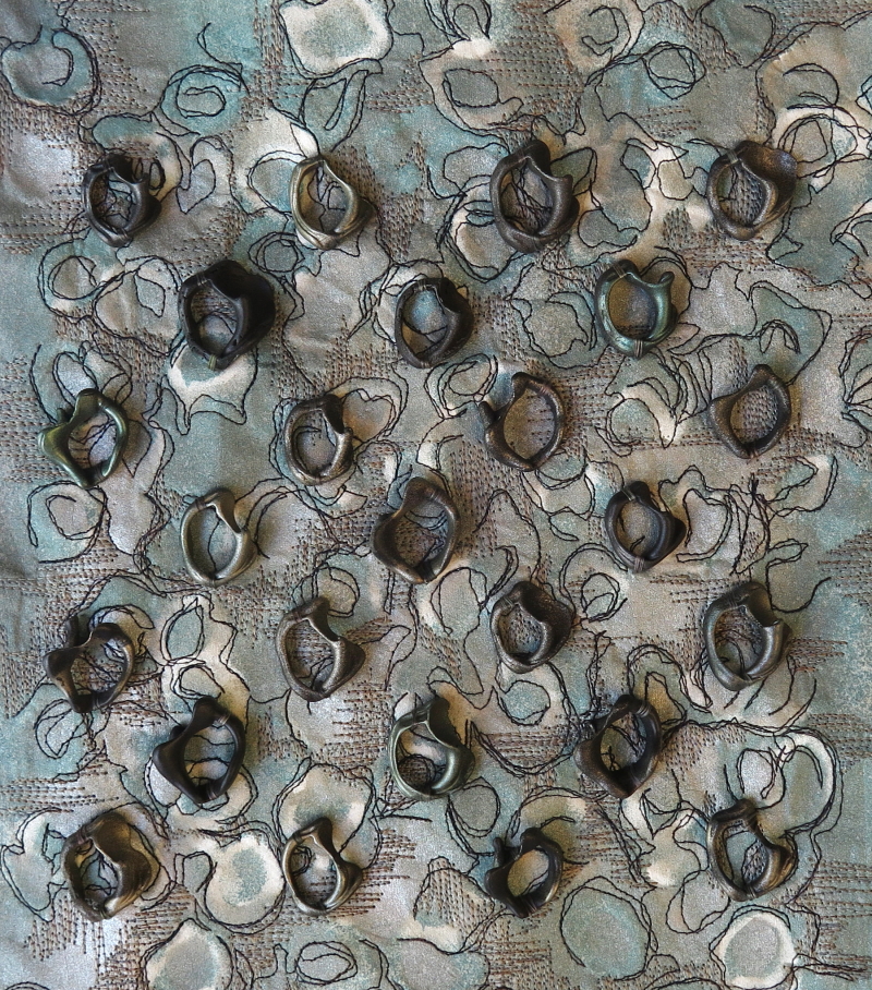 Dark shells uniformly sewn in a diamond pattern, on a background of cream and soft green coloured Evolon. The piece also has a large number of horizontal and vertical lines of light grey machine stitching as well as swirling black machine stitching, creating a feeling of movement.