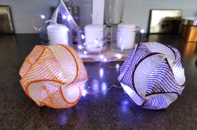 Two ball like structures of interwoven embroidered strips of Lutradur. One with orange and the other with purple stitching. Lit with small battery operated lights.