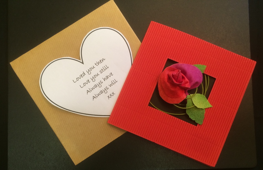 A square red corrugated card with a square aperture in the middle out of which an Evolon rose has been placed, attached to a spiral of thin green garden wire and three green leaves. To the left of the card is a brown paper envelope with a white heart bearing the words 'Loved you then, Love you still, Always have, Always will XXX' 