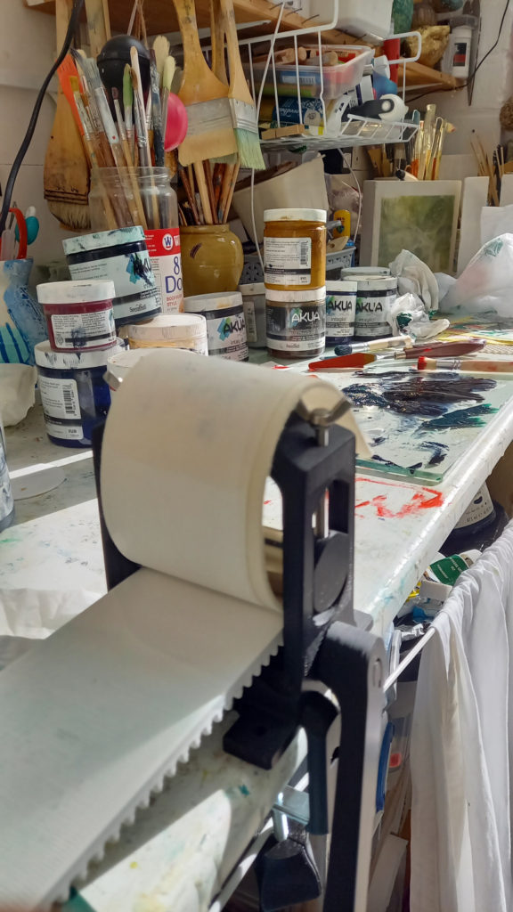An artist's workstation, covered with pots of paint and jars of paintbrushes. More paintbrushes hang from shelves above. In the foreground is a Mini 3D Etching Press.