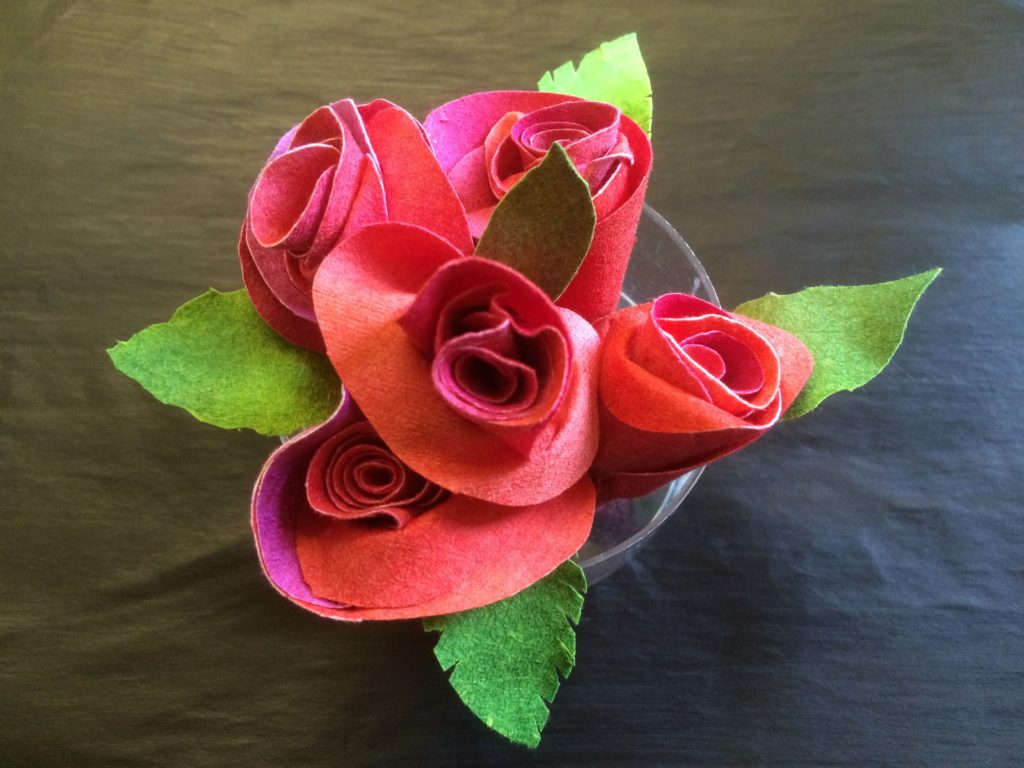 A ring of roses! A cluster of 5 red rose heads, positioned in a small clear dish and surrounded by green leaves.