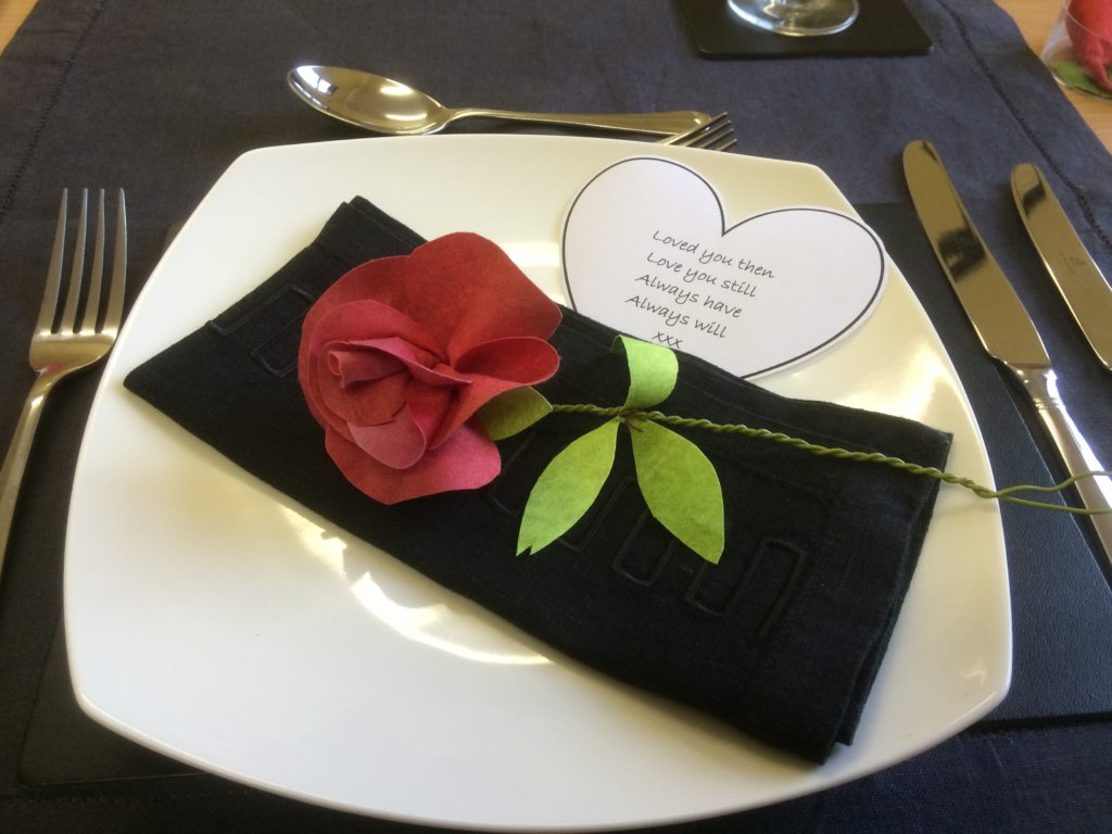 A single table setting on a dark blue tablecloth, consisting of a square white dinner plate, surrounded by cutlery. Resting diagonally on the plate is a single stem red rose placed on top of a folded black serviette. On the top righthand side of the plate is a heart shaped note, which reads - Loved you then, Love you still, Always have, Always will. XXX