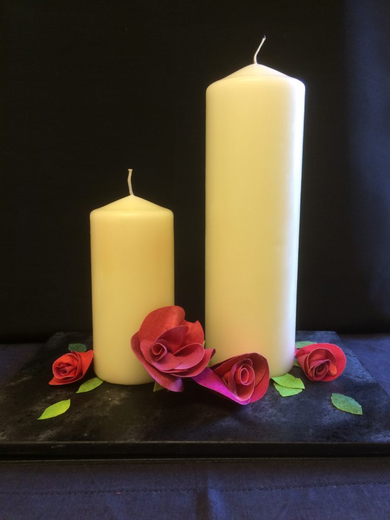 Two large cream candles. The candle on the right is almost twice the height of the candle on the left. They are unlit and standing against a dark blue background and table covering. Scattered around the base of the candles are four red roses and several individual green leaves, which have all been made from Evolon.