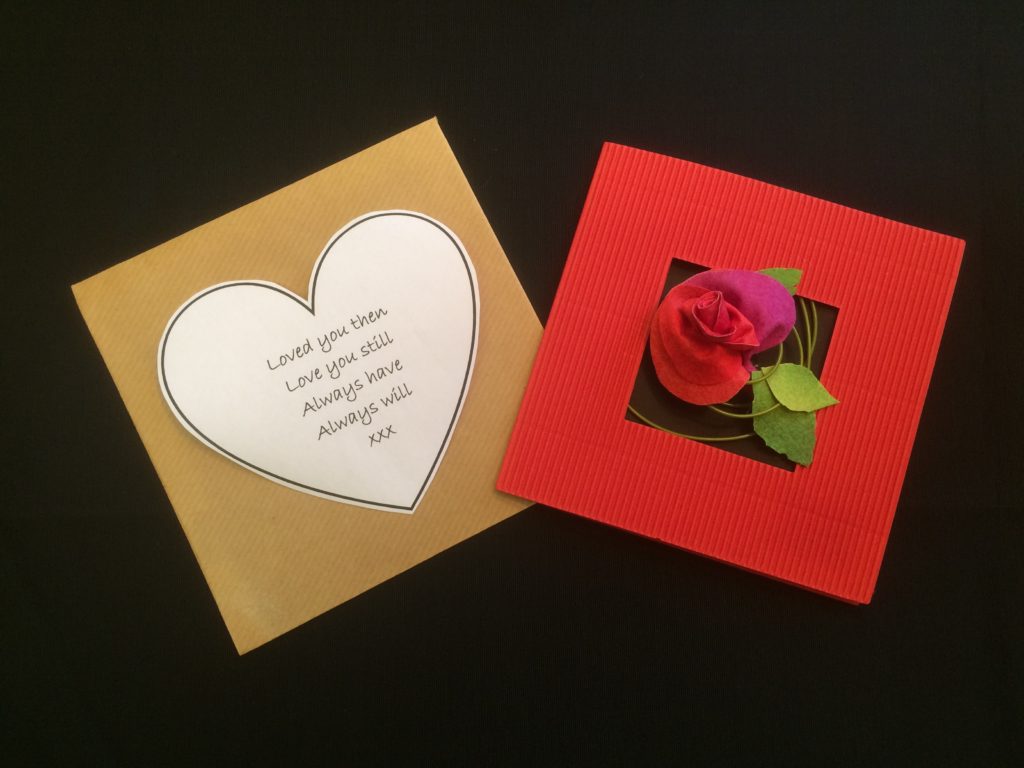 A square bright red Valentine card with a square hole in the centre, through which a single rose bud is emerging against a black background. The rose has been secured to the inside of the card with a length of thin green garden wire which has been wound round several times to form a circular pattern. Three green leaves also decorate the card. To the left of the card is a square brown envelope with a heart shaped note, which reads - Loved you then, Love you still, Always have, Always will. XXX