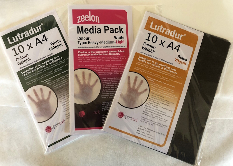 A photograph of three A4 packs of non-woven fabric, Lutradur 130gsm, Zeelon and Lutradur 70gsm Black.