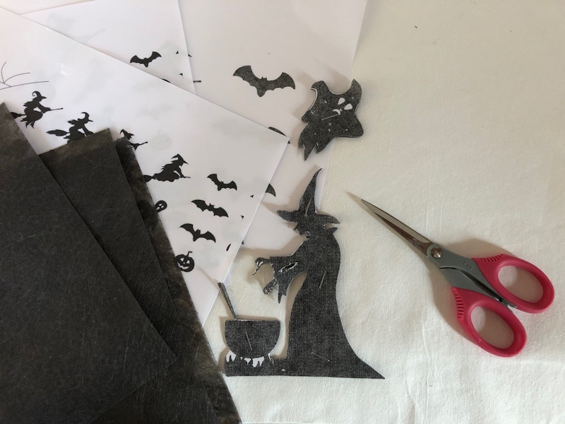 An image of a small black witch next to a cauldron, which has been cut out of a sheet of Lutradur 70gsm Black. There is also a small black ghost and black bat in the photograph. A small pair of scissors with red handles lie to the right.