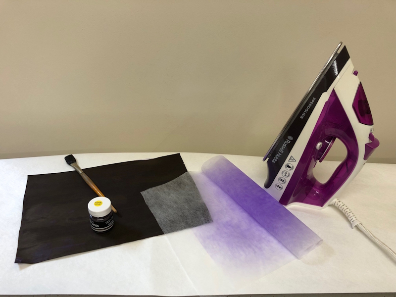 A picture of an iron, standing upright on an ironing board. On the board is a piece of lightly folded Zeelon Medium which has been coloured in a light violet shade, using transfer paint. A painted sheet of A4 paper is also on the ironing board, together with a small bottle of paint and a paintbrush.