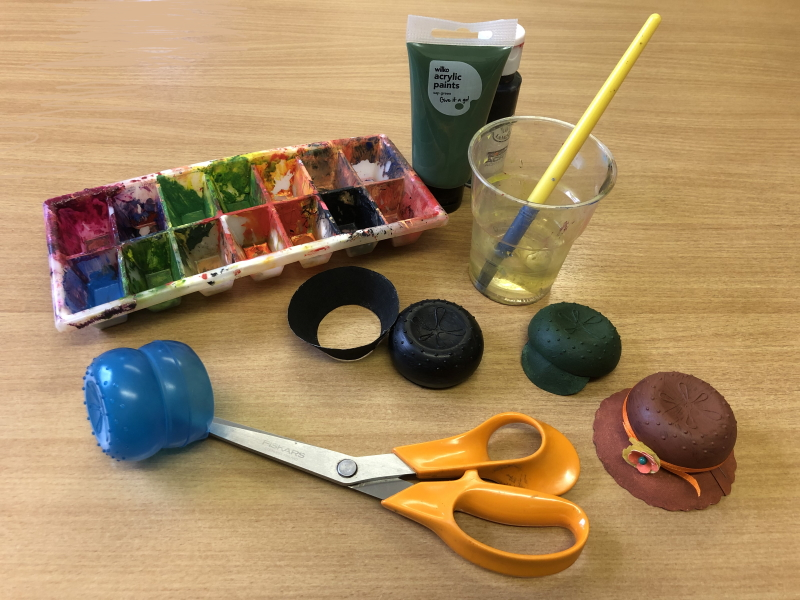 A blue liquid soap dispenser (the type used in automatic washing machines), a pair of large scissors with orange handles, pictured with some tubes of green and black acrylic paint, paintbrush standing in a plastic cup of water, mixing tray, plus the little green cap, the hat with the flower and the bowler with the crown and rim shown separately.

