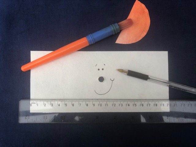 A white rectangle of Evolon with a small hole cut in the centre, (in the position of the nose) surrounded by two little black eyes and a smile. Also a small semicircle of orange painted Evolon, a paintbrush, black biro and a clear plastic ruler showing the measurement of the Evolon.
