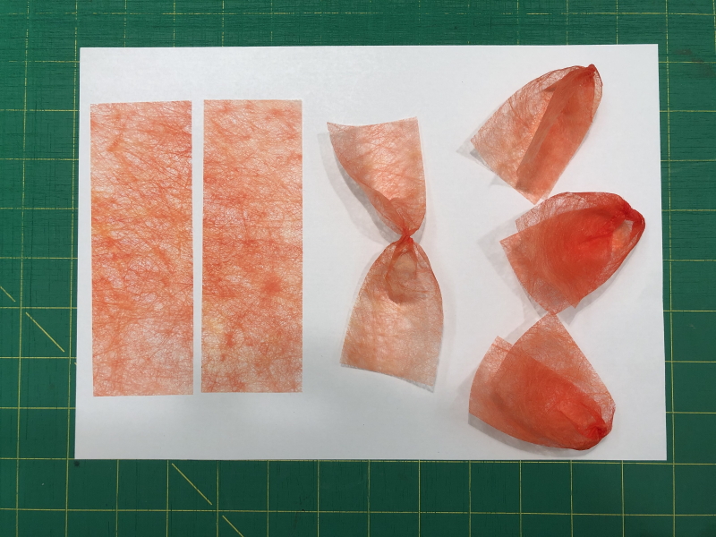 Six pieces of orange coloured Lutradur all the same size. Two rectangular strips lie on the left of the photograph. In the centre is a single strip which has been twisted in the middle and on the right are three 'petals', formed by folding the twisted strips of Lutradur over on themselves. 