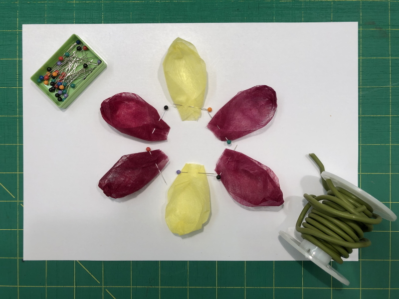A white A4 piece of paper resting on a cutting board. In the centre are six petals, lying in a flower formation, 2 yellow and 4 wine red.  A small green box of coloured pins is positioned in the top left corner. All the lower corners of the petals have been folded over and pinned, ready for sewing, to give the desired spoon shape. In the bottom right hand corner of the photograph can be seen a roll of  plastic coated flexible garden wire. 