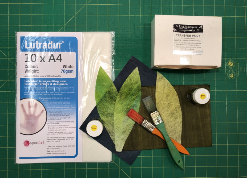 An A4 pack of Lutradur 70gsm, lying on a green measuring board. In the top right hand corner is a white box of transfer paints and in the lower right hand corner are two paintbrushes, lying on a couple of sheets of green transfer painted paper. There are also 3 green leaves cut from Lutradur 70gsm, shaped to resemble those of a tulip.