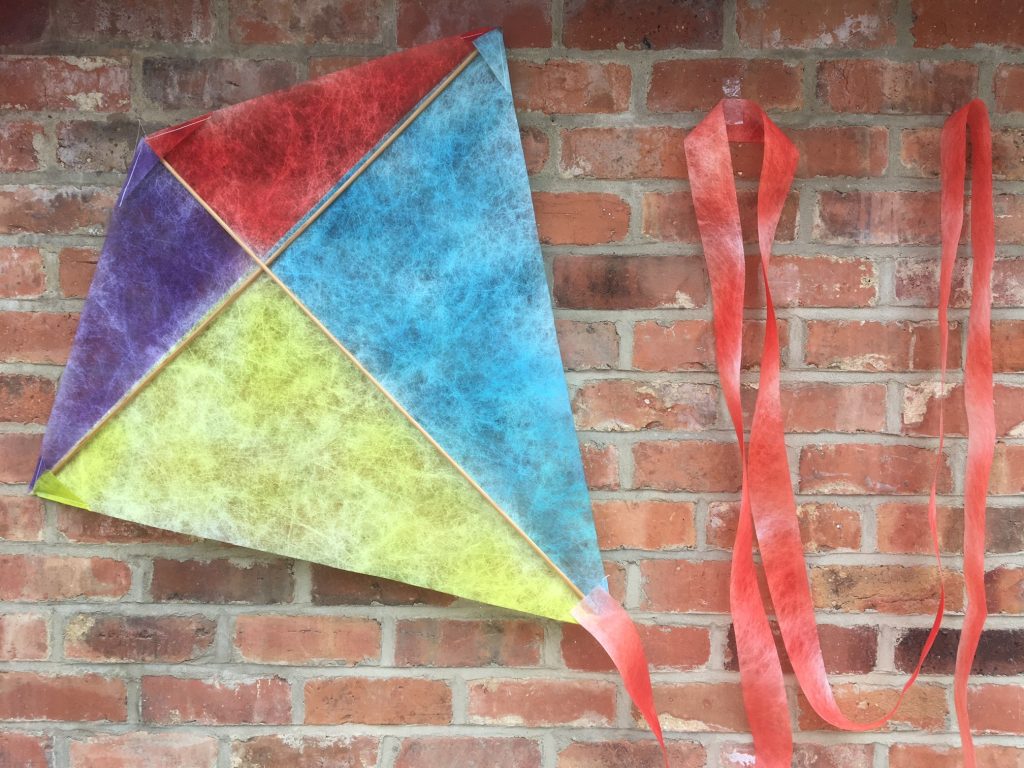 A diamond shaped kite made from Lutradur hanging on a rustic brick wall. The kite has a long red tail which is looped and hung to the right of the kite. The kite is painted in 4 blocks of colour. The top two triangular sections are purple and red and the bottom two are yellow and turquoise. The kite is kept taut by two dowelling rods which brace the kite from top to bottom and left to right.