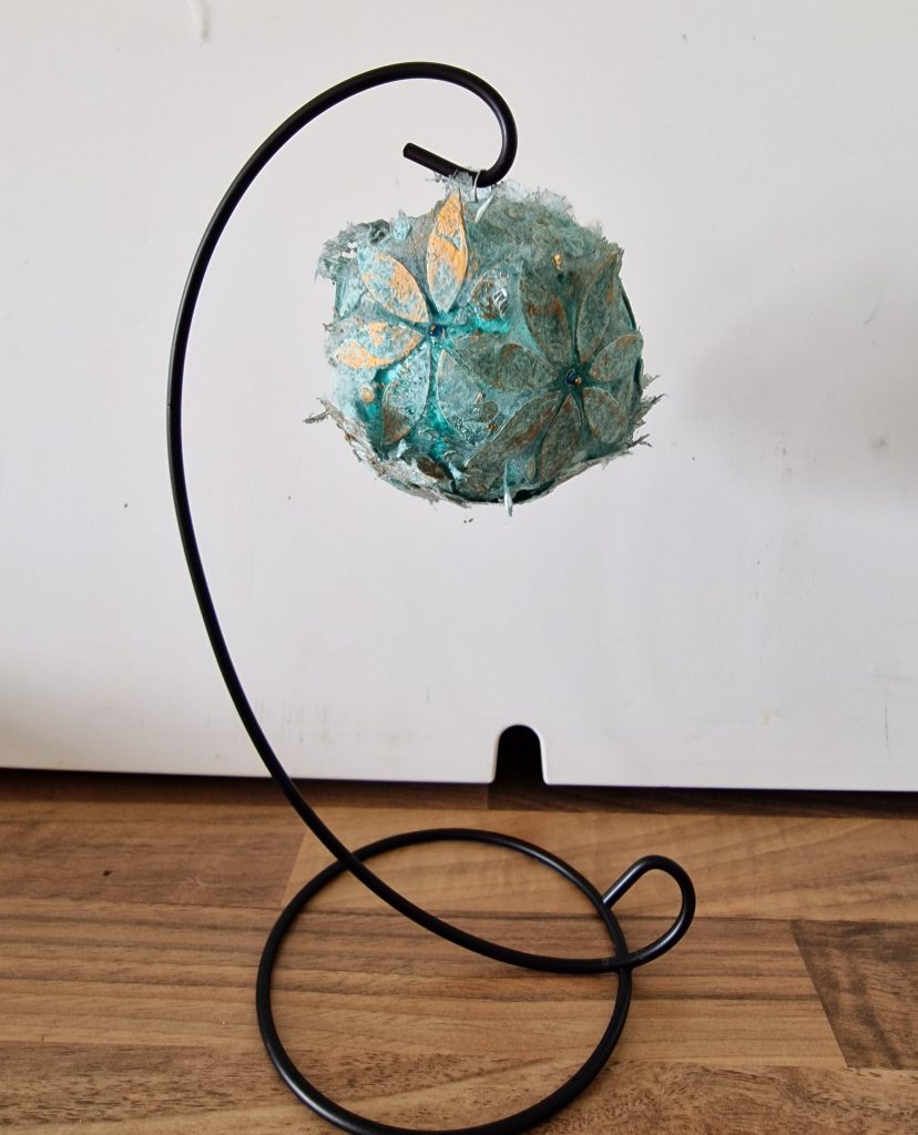 A round bauble covered with pale green flowers cut from Lutradur. Decorated with shimmering paint and gold beads.