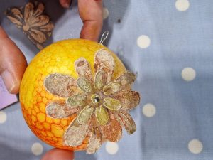 A brightly painted orange and yellow bauble, showing a flower with two layers of petals and a gold bead in the centre.