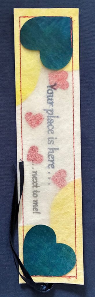 A white rectangular bookmark with red machine stitching all round the edge. The text on the bookmark reads 'Your place is here . . . next to me!' The top layer of the bookmark is semi transparent and inside can be seen half a dozen small shiny hearts, which are free to move about. In diagonal corners are two mid blue hearts, attached to the front of the bookmark.