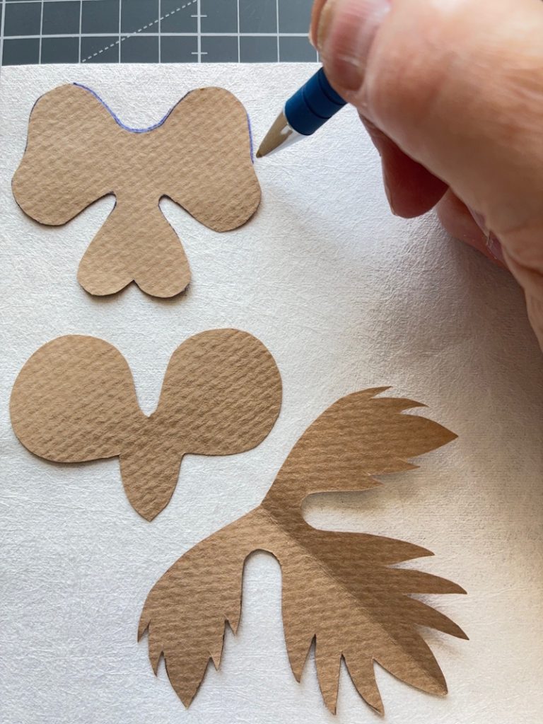 A close-up image showing a hand holding a blue pen, drawing around cut-out shapes of pansy petals and leaves on a piece of Evolon.
