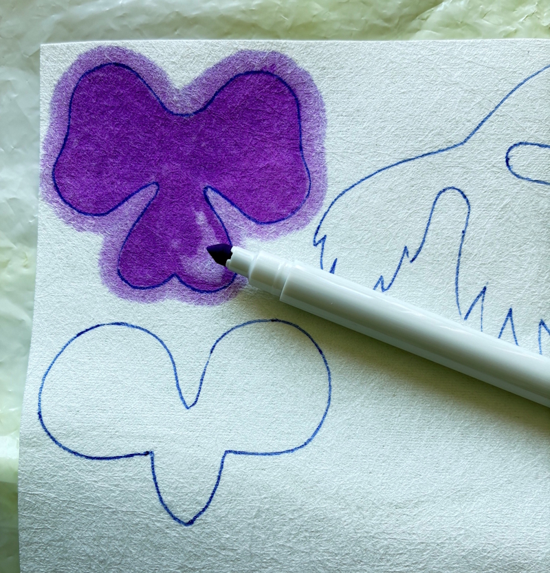 A Crayola fibre tip pen is placed next to two drawn flower shapes. One flower shape has been almost completely coloured j 
in with a purple hue. The ink is shown to be spreading beyond the outline of the petals.