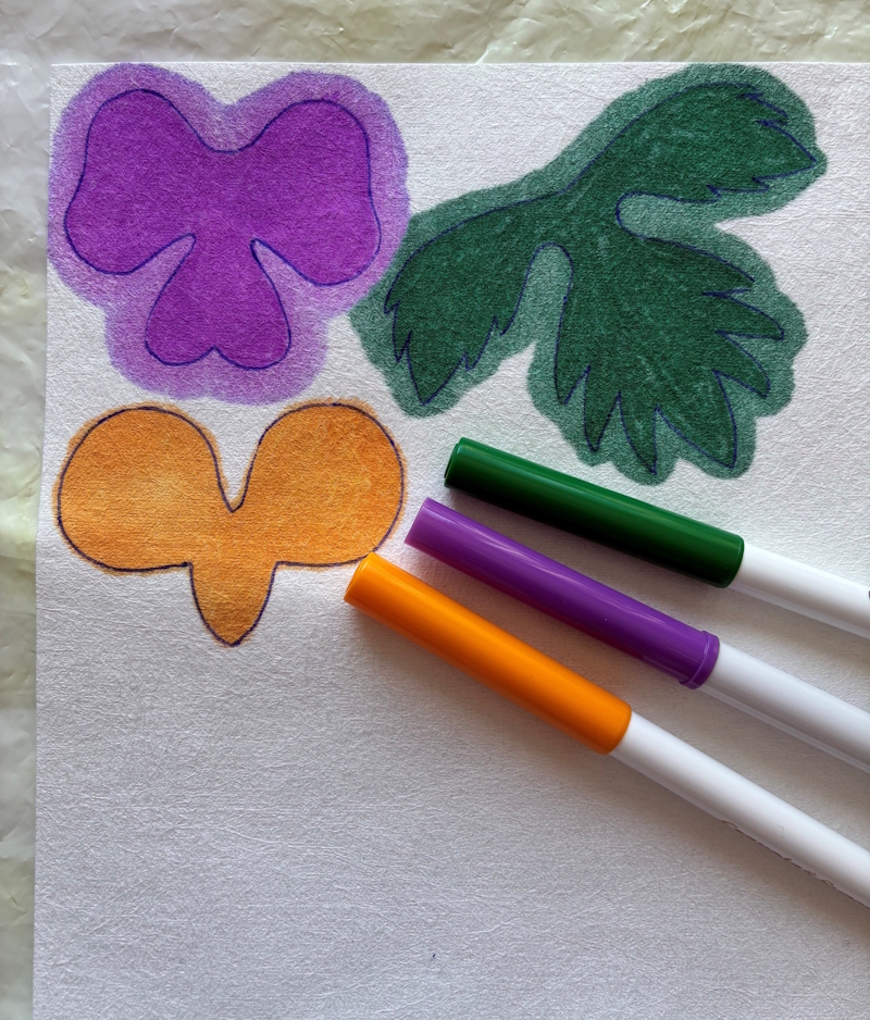Two petal shapes coloured in by fibre pens, one solid purple and the other orange, alongside some dark green leaves. Three corresponding fibre tip pens are lying next to the shapes.