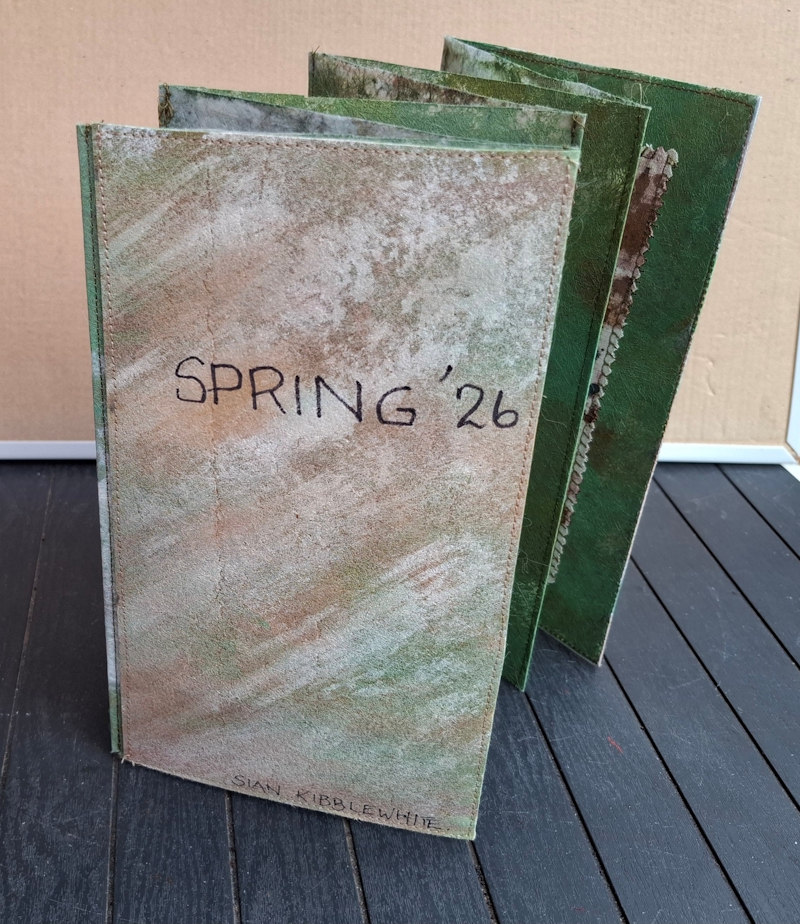 A folded concertina style display booklet, consisting of 8 sides, includng the front and back pages. The words  'Spring '26' are written on the front and the name Sian Kibblewhite appears on the front right hand corner.. The inside pages are not clearly seen in this photograph, but the overall colours are muted earthly tones.